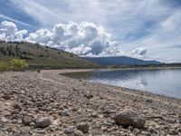 a lake with rocks on the shore and mountains in the background along side it and snow capped hills