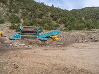 two yellow bulldozers work on sand and gravel in a mountain valley while another one looks at it