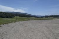 a dirt field and road near pine trees and mountain tops against blue sky with wispy clouds