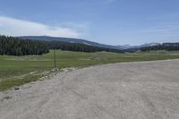 a dirt field and road near pine trees and mountain tops against blue sky with wispy clouds