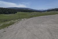 a dirt field and road near pine trees and mountain tops against blue sky with wispy clouds