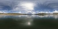 a pond surrounded by water with some clouds above it and the sun reflecting off the water and mountains in the background