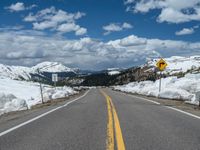 snow covers the roadway and snowy mountains on a sunny day, with a yellow warning sign in front of it