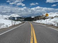 snow covers the roadway and snowy mountains on a sunny day, with a yellow warning sign in front of it