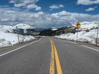 snow covers the roadway and snowy mountains on a sunny day, with a yellow warning sign in front of it