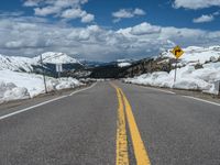 snow covers the roadway and snowy mountains on a sunny day, with a yellow warning sign in front of it