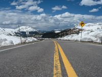 snow covers the roadway and snowy mountains on a sunny day, with a yellow warning sign in front of it