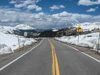 snow covers the roadway and snowy mountains on a sunny day, with a yellow warning sign in front of it