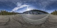 a curved road and some snowy mountains and trees in the background with a cloudy sky above it