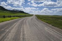 a dirt road leading to an empty fence and grassy field with animals in it, on a cloudy day