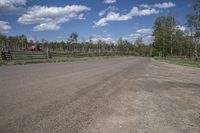 an empty dirt road in front of wooden fences and clouds overhead a large tree lined fence is at the edge of the street