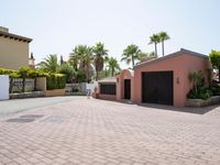 an driveway of many colors that is bordered by palm trees and brick tiles, and some small plants in front of the garage