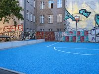 a blue basketball court near some brown buildings with colorful graffiti walls on it and blue floor