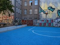 a blue basketball court near some brown buildings with colorful graffiti walls on it and blue floor