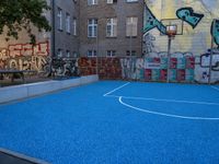 a blue basketball court near some brown buildings with colorful graffiti walls on it and blue floor