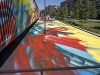 a painting with colorful colors is on the floor of a train carriage with a large metal container near a line of buildings in the city