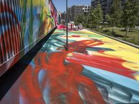 a painting with colorful colors is on the floor of a train carriage with a large metal container near a line of buildings in the city