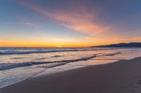 a colorful sunset at the beach with waves rolling in towards the shore and a plane flying through