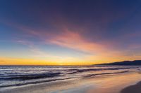 a colorful sunset at the beach with waves rolling in towards the shore and a plane flying through