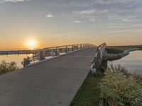 bridge leading to the beach near water and sun sets over ocean grass and bushes in front