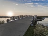 bridge leading to the beach near water and sun sets over ocean grass and bushes in front