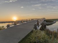 bridge leading to the beach near water and sun sets over ocean grass and bushes in front