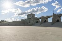 a concrete bridge spanning the width of a skateboard park and ramps under the sun and clouds