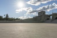 a concrete bridge spanning the width of a skateboard park and ramps under the sun and clouds