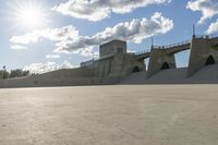 a concrete bridge spanning the width of a skateboard park and ramps under the sun and clouds