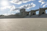 a concrete bridge spanning the width of a skateboard park and ramps under the sun and clouds