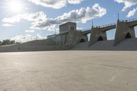 a concrete bridge spanning the width of a skateboard park and ramps under the sun and clouds