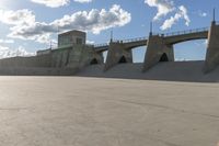 a concrete bridge spanning the width of a skateboard park and ramps under the sun and clouds