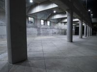 a concrete building has pillars and lights lit up and outside it at night, and there is a person with a skateboard inside