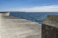 a large concrete fence along the water's edge with sculpture in distance on a clear day