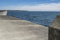 a large concrete fence along the water's edge with sculpture in distance on a clear day