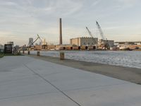 a concrete pathway next to a body of water under construction next to an industrial area with large cranes