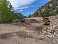 two yellow bulldozers work on sand and gravel in a mountain valley while another one looks at it