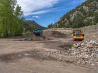 two yellow bulldozers work on sand and gravel in a mountain valley while another one looks at it