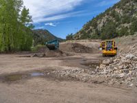 two yellow bulldozers work on sand and gravel in a mountain valley while another one looks at it