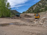 two yellow bulldozers work on sand and gravel in a mountain valley while another one looks at it