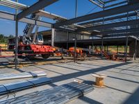 a red vehicle driving in an open floor building under construction, with a worker on a fork lifting a large beam to put metal in place