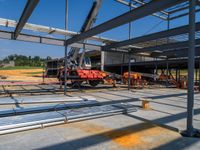 a red vehicle driving in an open floor building under construction, with a worker on a fork lifting a large beam to put metal in place