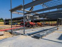 a red vehicle driving in an open floor building under construction, with a worker on a fork lifting a large beam to put metal in place