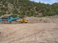 two yellow bulldozers work on sand and gravel in a mountain valley while another one looks at it