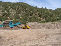 two yellow bulldozers work on sand and gravel in a mountain valley while another one looks at it