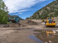 two yellow bulldozers work on sand and gravel in a mountain valley while another one looks at it