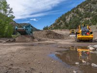 two yellow bulldozers work on sand and gravel in a mountain valley while another one looks at it