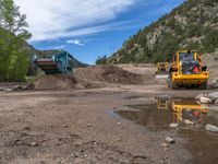 two yellow bulldozers work on sand and gravel in a mountain valley while another one looks at it