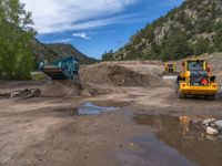 two yellow bulldozers work on sand and gravel in a mountain valley while another one looks at it