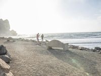 a couple are walking down the beach towards the ocean with rocks, boulders and benches